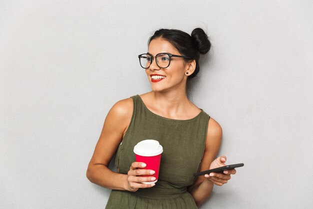 Portrait of a smiling young woman isolated, using mobile phone, holding takeaway coffee cup