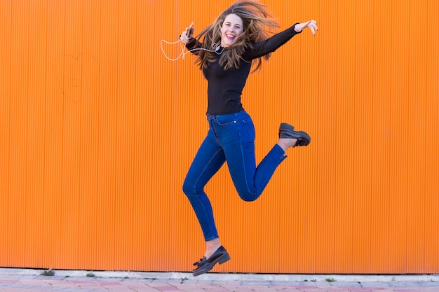 Photo portrait of smiling young woman listening music on mobile phone while jumping against orange wall