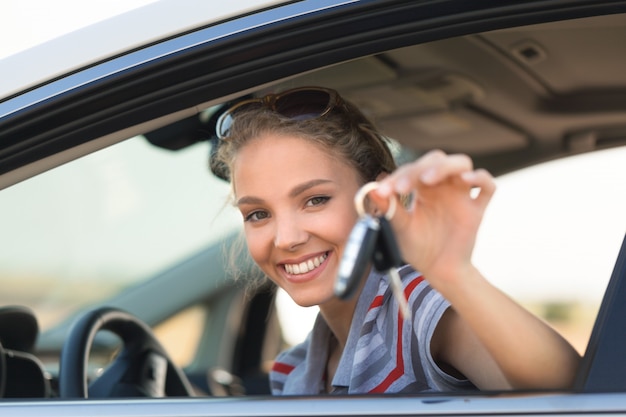 車のキーを示す彼女の車の窓の外を見ている笑顔の若い女性の肖像画