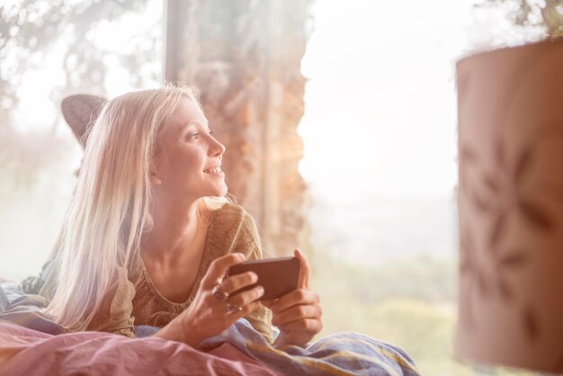 Portrait of smiling young woman lying on bed with cell phone looking through window