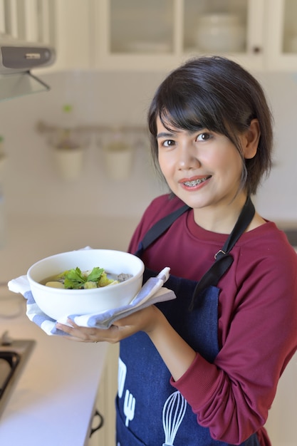 Photo portrait of smiling young woman in modern kitchen and holding a bowl of soup with tofu