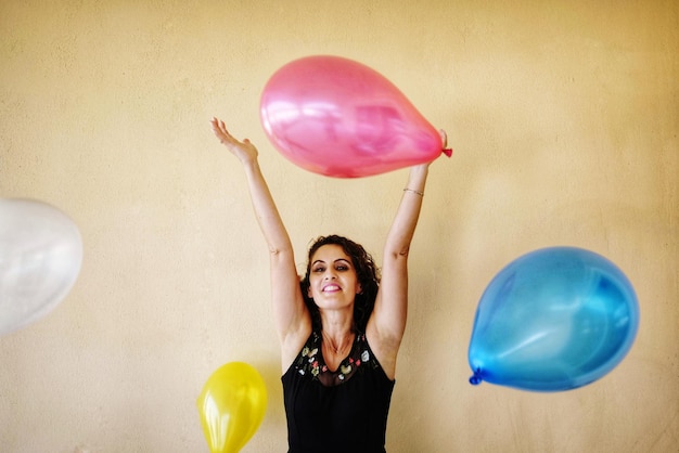 Photo portrait of smiling young woman playing with balloons while standing against wall