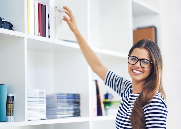 Portrait of a smiling young woman reaching for a book on a shelf at home