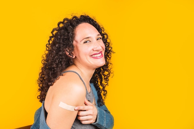 Photo portrait of a smiling young woman showing arm after coronavirus vaccination - sitting in chair with dipped sleeve on yellow background.