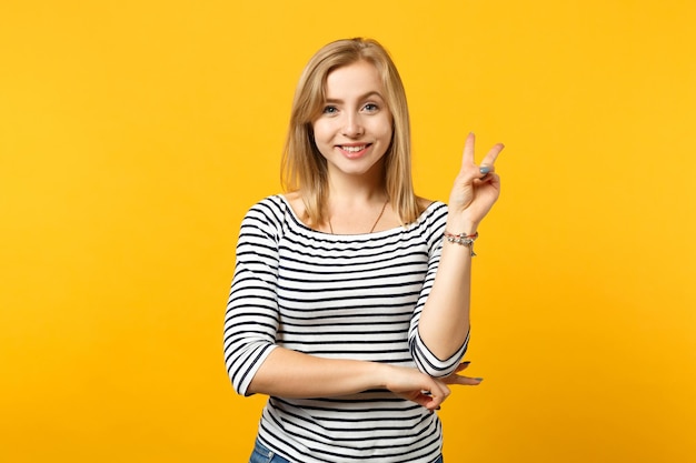 Portrait of smiling young woman in striped clothes looking camera, showing victory sign isolated on yellow orange background in studio. People sincere emotions, lifestyle concept. Mock up copy space.