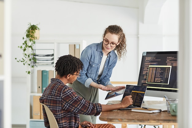 Portrait of smiling young woman talking to African-American man writing code while working in web development studio, copy space