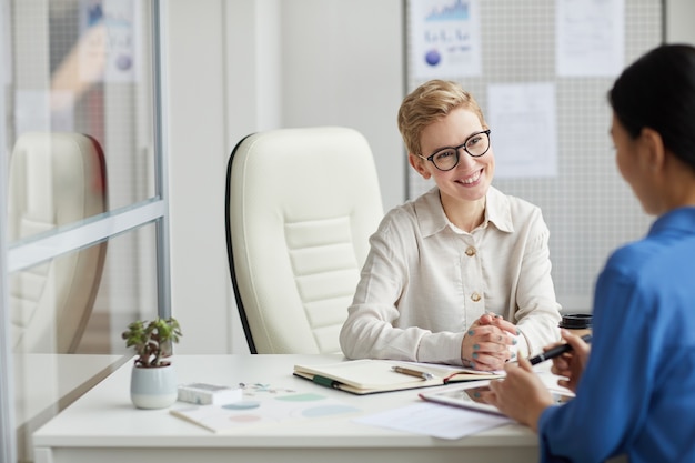 Portrait of smiling young woman talking to client or partner while working at desk in office cubicle, copy space