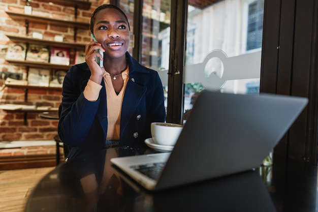 Portrait of a smiling young woman using laptop and talking on mobile phone in coffeeshop