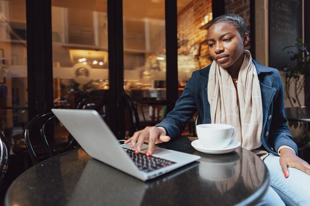 Портрет улыбающейся молодой женщины, использующей ноутбук и разговаривающей по мобильному телефону в кафе