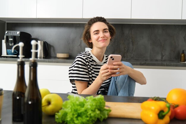 Photo portrait of smiling young woman using mobile phone in kitchen