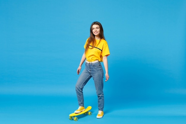 Ritratto di giovane donna sorridente in abiti casual vivaci che guarda la fotocamera e in piedi con lo skateboard giallo isolato su sfondo blu della parete in studio. concetto di stile di vita delle persone. mock up spazio di copia.