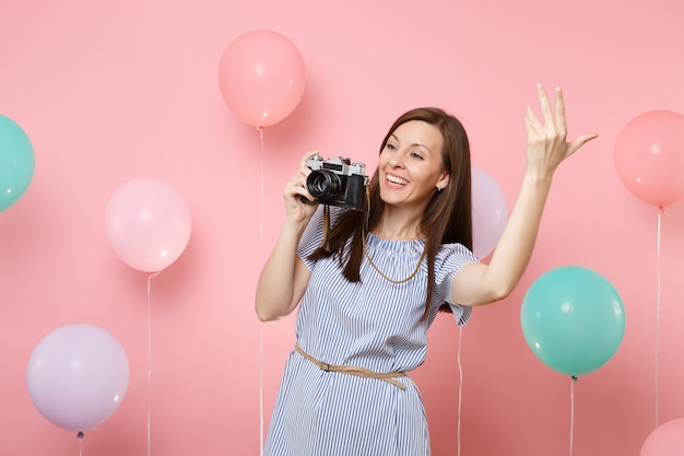 Ritratto di giovane donna sorridente che indossa abito blu che tiene retro macchina fotografica d'epoca agitando la mano su sfondo rosa con mongolfiere colorate. festa di compleanno persone sincere emozioni concetto.