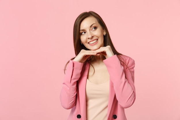 Portrait of smiling young woman wearing jacket put hand prop up on chin looking aside isolated on pastel pink wall background in studio. People sincere emotions, lifestyle concept. Mock up copy space.