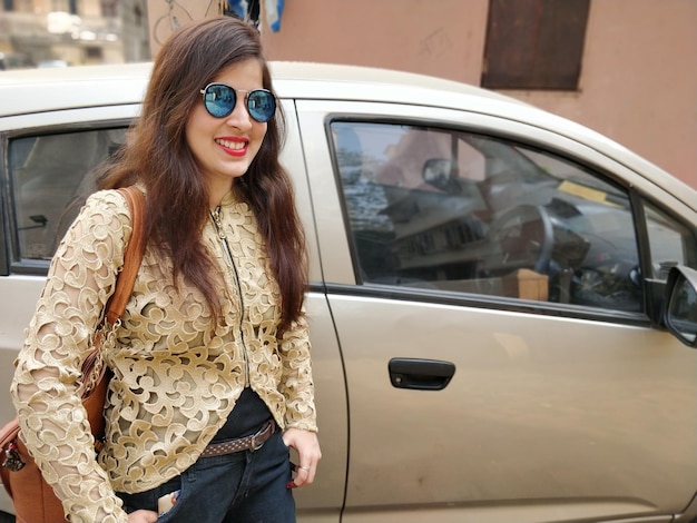 Portrait of smiling young woman wearing sunglasses standing against car