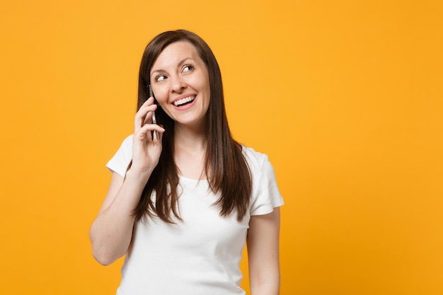 Portrait of smiling young woman in white casual clothes looking up, talking on mobile phone isolated on bright yellow orange wall background in studio. People lifestyle concept. Mock up copy space.