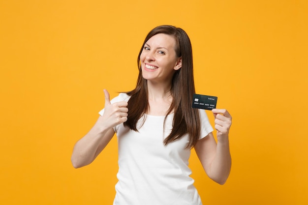 Portrait of smiling young woman in white casual clothes showing thumb up, holding credit bank card isolated on yellow orange wall background in studio. People lifestyle concept. Mock up copy space.