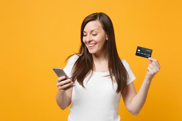 Portrait of smiling young woman in white casual clothes using mobile phone, holding credit bank card isolated on yellow orange wall background in studio. people lifestyle concept. mock up copy space