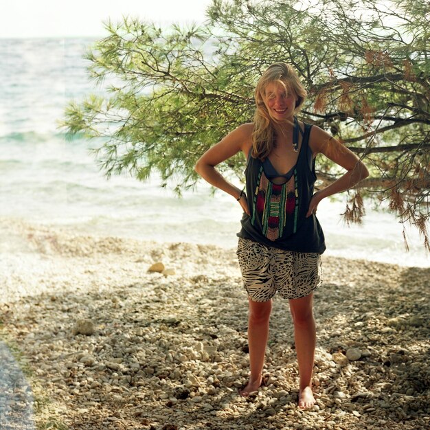 Portrait of smiling young woman with hands on hip standing against tree