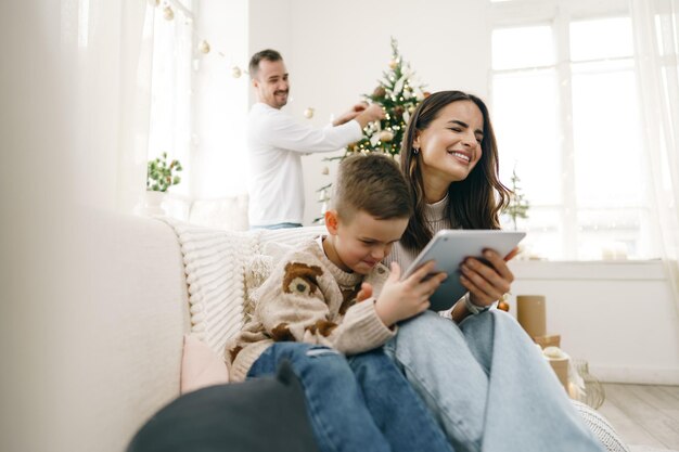 Портрет улыбающейся молодой женщины с сыном с помощью цифрового планшета на зимних каникулах дома