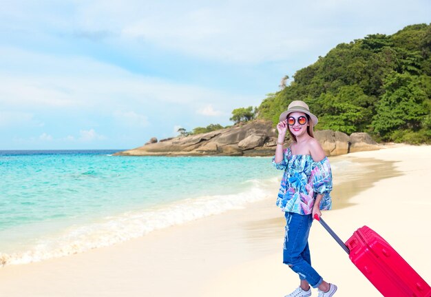 Portrait of smiling young woman with suitcase at beach against sky