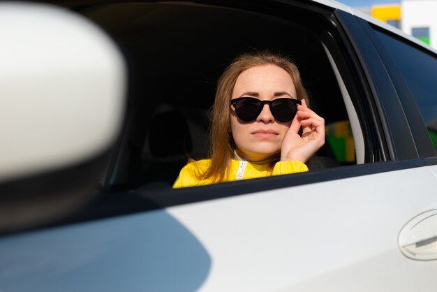 Ritratto di giovane donna sorridente in una giacca gialla che abbassa gli occhiali da sole seduta nella sua auto.