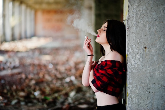 Portrait smoking girl with red lips wearing a red checkered shirt with bare shoulders posed sexy background abadoned place