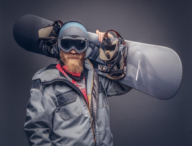 Portrait of a snowboarder dressed in a full protective gear for extream snowboarding posing with a snowboard on his shoulder at a studio. Isolated on a gray background.