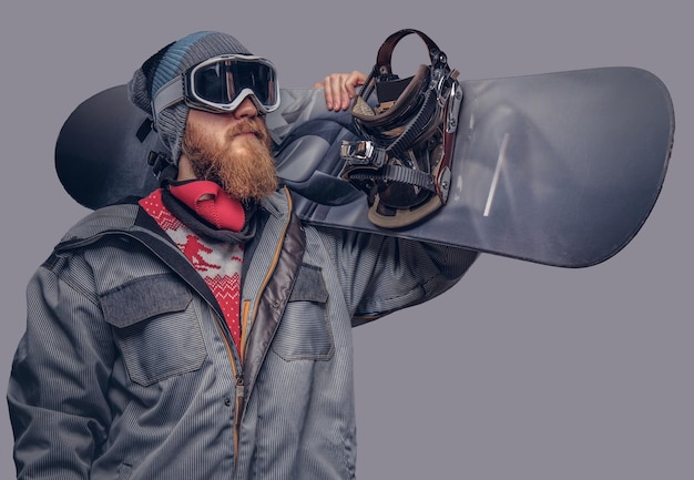 Portrait of a snowboarder dressed in a full protective gear for extream snowboarding posing with a snowboard on his shoulder at a studio. Isolated on a gray background.