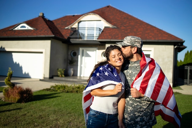 Foto ritratto di soldato in uniforme che bacia sua moglie e tiene la bandiera americana davanti alla loro casa