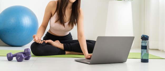 운동복을 입은 아시아 미인 몸매 슬림 여성이 앉아 휴식을 취하고 요가를 하는 소녀와 가정의 침실에서 노트북 컴퓨터로 피트니스 운동을 합니다.다이어트 개념피트니스 및 건강