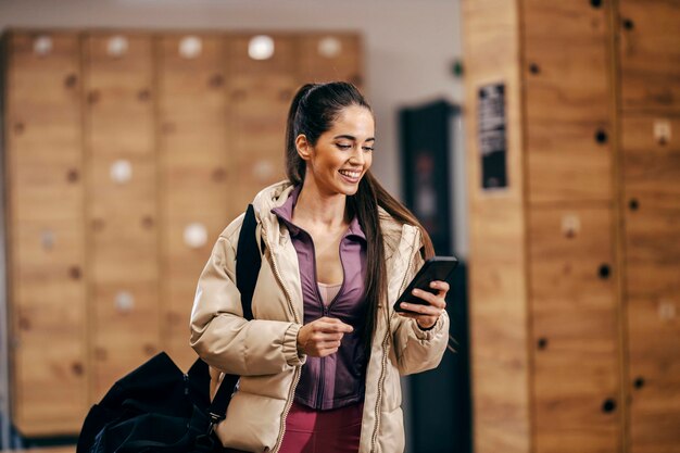 Portrait of a sportswoman standing in fitting room with her sports bag and looking at the phone