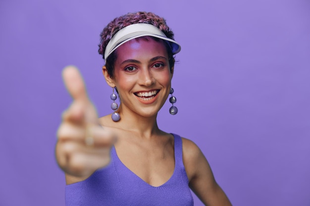 Portrait of a sporty fashion woman posing smiling with teeth and pointing a finger at the camera in a purple yoga tracksuit and a transparent cap on a purple monochrome background high quality photo