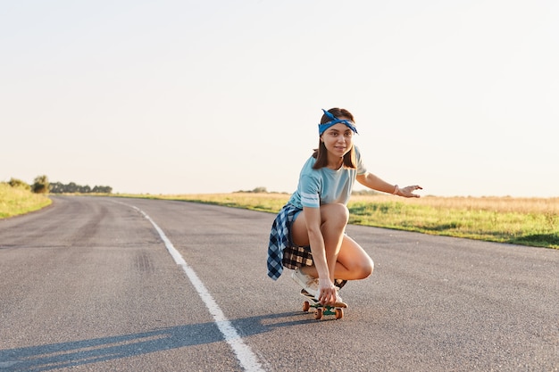 Портрет спортивной женщины в повседневной одежде, сидящей на корточках на скейтборде, смотрящей вдаль с концентрированным выражением лица, наслаждающейся скейтбордингом, здоровым образом жизни.