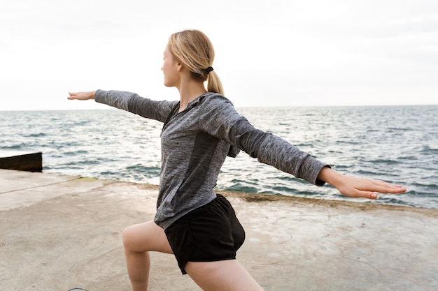 Portrait of sporty woman wearing tracksuit doing exercise while working out on pier near seaside in morning