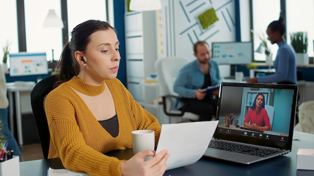 Portrait of startup employee presenting paper with charts using laptop in casual video call conference with manager sitting at desk. Relaxed woman in busy department talking with executive.