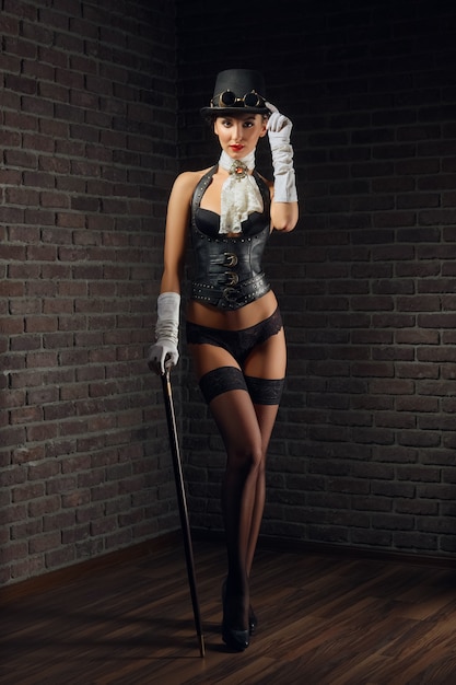 Portrait of steampunk girl in hat with glasses and cane in leather vest and stockings.