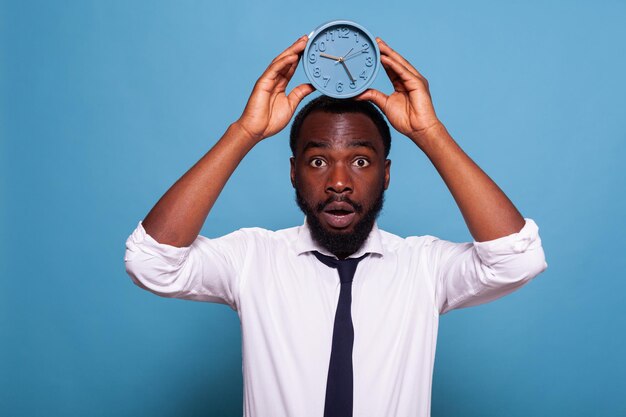 Photo portrait of stressed office worker holding alarm clock over head showing passing time concept on blue background. anxious businessman in white shirt and tie looking nervous feeling deadline pressure.