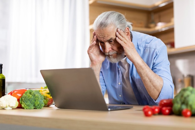 Portrait of stressed senior man using laptop in kitchen interior