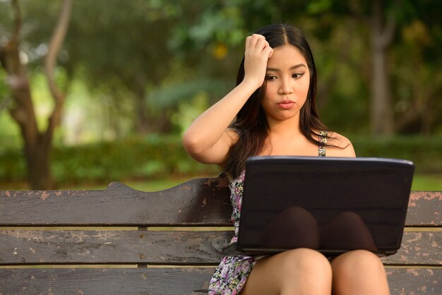 Портрет подчеркнутой молодой азиатской женщины, использующей ноутбук и потрясенной, глядя на парк