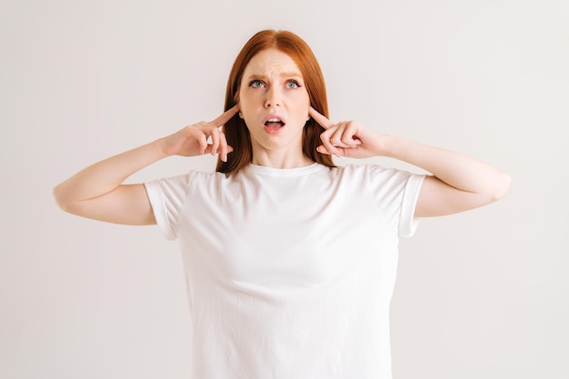 Portrait of stressed young woman covering her ears and gesticulating say no blablabla standing on white isolated background in studio Irritated redhead female showing nonsense content