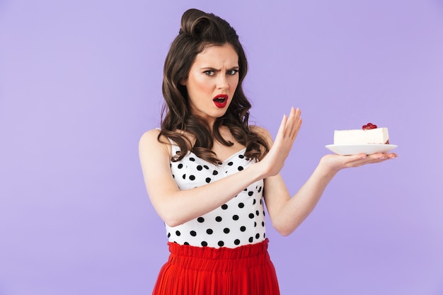 Portrait of strict pin-up woman in vintage polka dot dress holding and rejecting sweet cheesecake isolated over violet wall