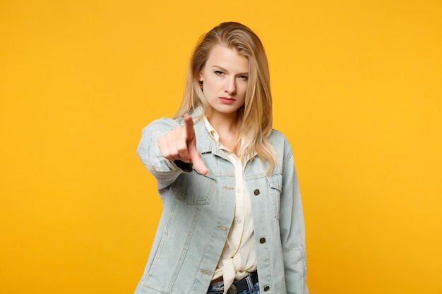 Portrait of strict serious young woman in denim casual clothes pointing index finger on camera isolated on bright yellow orange wall background in studio. people lifestyle concept. mock up copy space