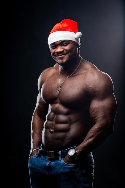 Portrait of a strong afroamerican man wearing Christmas hat showing off his physique against back background Man with perfect body posing at the studio while looking at the camera