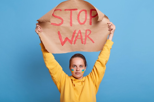 Portrait of strong brave woman in yellow hoodie with Ukrainian flag on cheeks protesting on demonstration showing poster with stop war words posing isolated over blue background