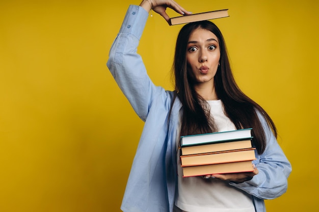 Portrait of student girl balancing books on head and hand while standing against yellow background