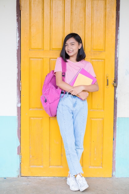 Portrait student young girl on yellow door background with pink backpacksmiling holding books wear pink shirt and jean