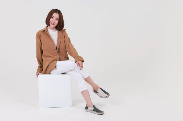 Portrait studio cutout shot of Asian young pretty short hair female model in long brown coat jacket casual shoes sitting crossed legs smiling look at camera on box chair posing on white background.