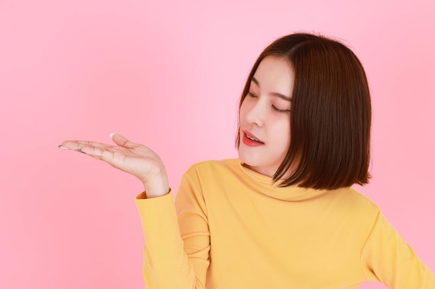 Portrait studio cutout shot of asian young pretty short hair\
female model in yellow long sleeve shirt stand smile to look at the\
camera and pointing finger on pink background.