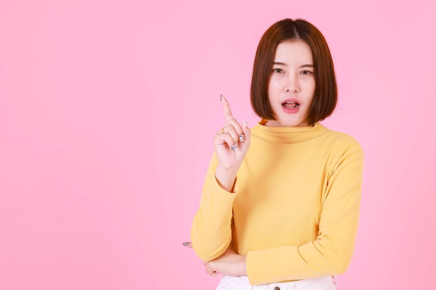 Ritratto studio ritaglio colpo di modello femminile asiatico giovane capelli piuttosto corti in camicia a maniche lunghe gialla stand sorriso a guardare la telecamera e puntare il dito su sfondo rosa.