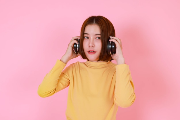 ピンクの背景で音楽の歌を聴きながら大きな黒いヘッドフォンを調整しながら立っている黄色の長袖シャツのアジアの若いかなり短い髪の女性モデルのポートレートスタジオカットアウトショット。